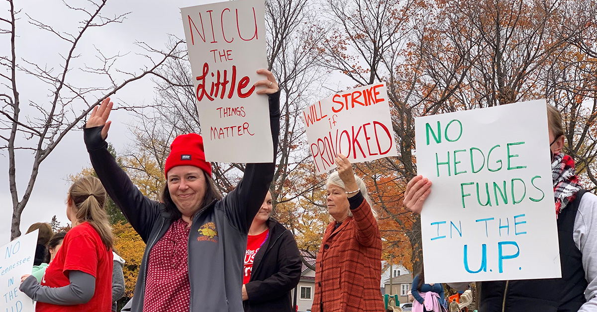 Group of nurses outside hold signs against greedy out-of-state hospital corporations