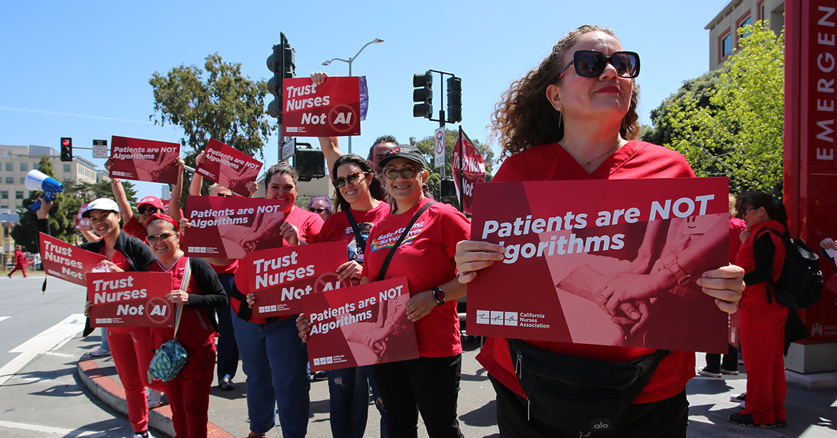 Group of nurses outside holding signs "Patients are Not Algorithms" and "Trust Nurses, Not A.I."