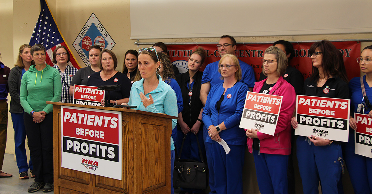 Group of nurse standing behind nurse at podium with "Patients Before Profits" signs