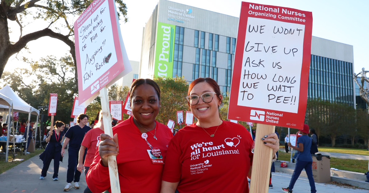 Two nurses outside hospital smiling and holding signs, "Stand up for something or fall for anything" and "We won't give up"