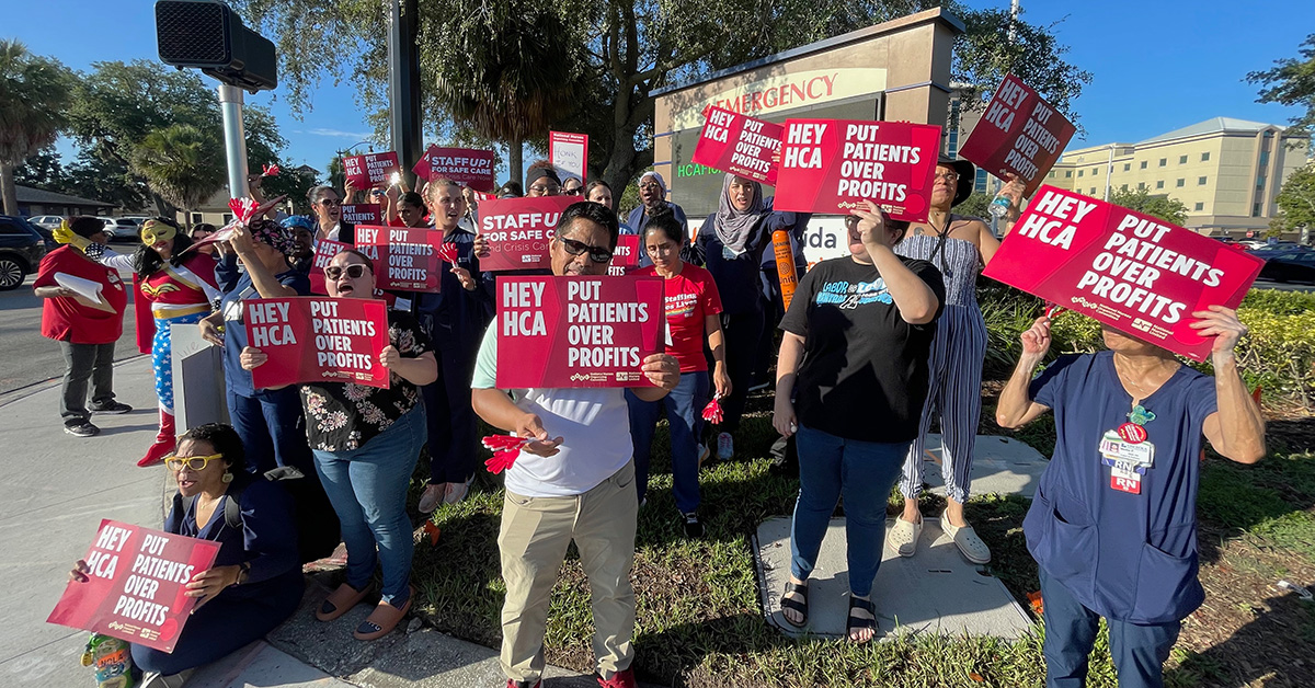 Large group of nurses outside hospital, holding signs "Hey HCA, Put Patients Over Profit"