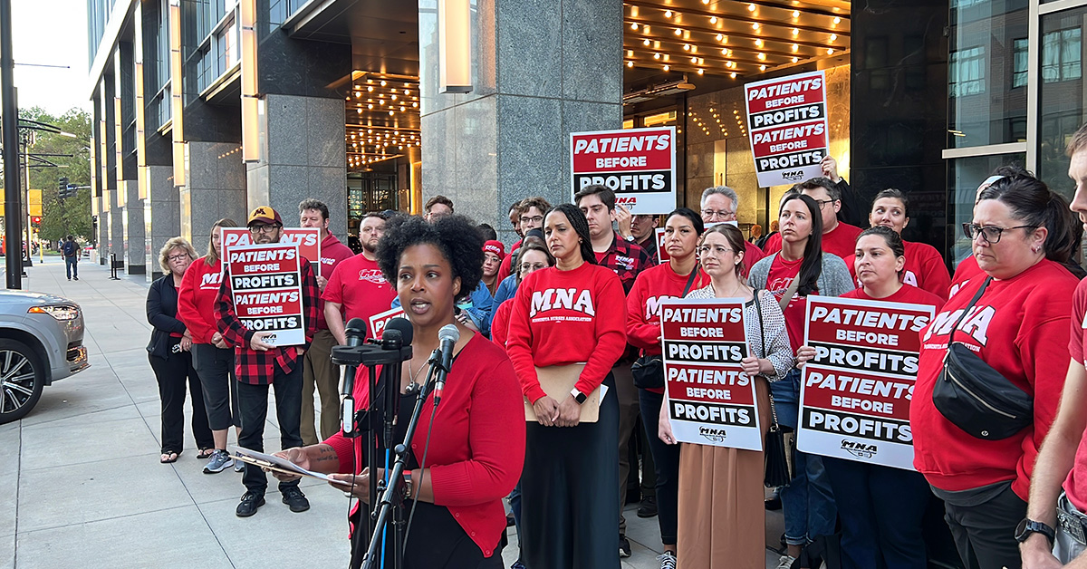 Nurse speaking at podium outside, more nurses behind her holding signs "Patients before profits"