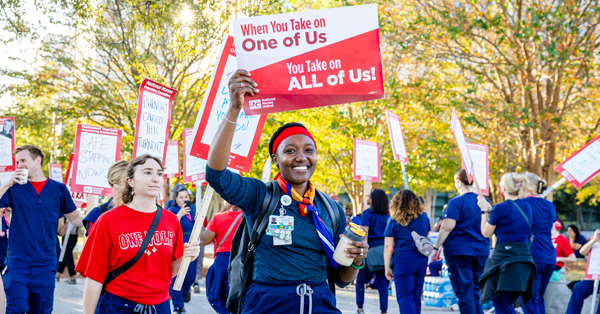 Nurse on picket line holding sign "When You Take On One Of Use, You Take On All Of Us"