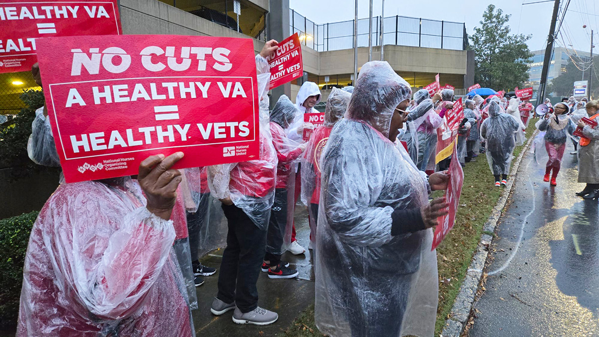 Nurses on picket line outside hospital in the rain. One holds sign "No Cuts: A Healthy VA = Healthy Vets