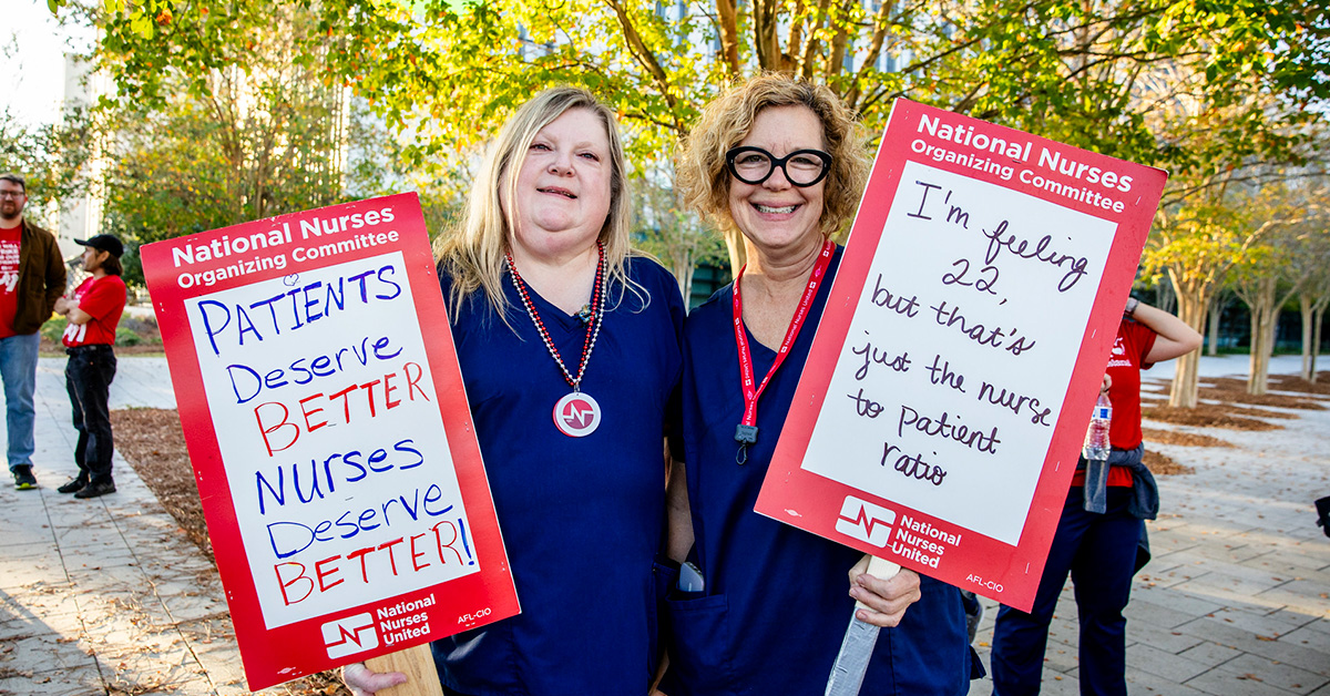 Two nurses smiling, holding signs "Patients deserve better, nurses deserve better"