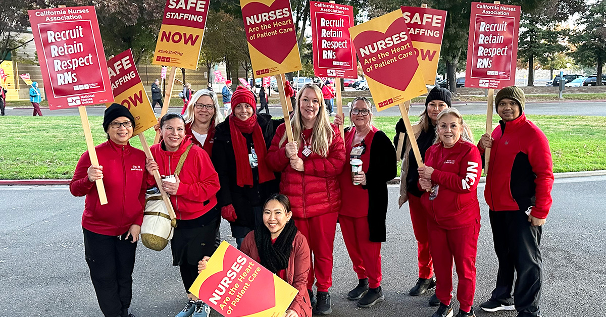 Group of nurses outside holding signs "Safe Staffing NOW", "Recruit, Retain, Respect RNs", and "Nurses are the heart of patient care"