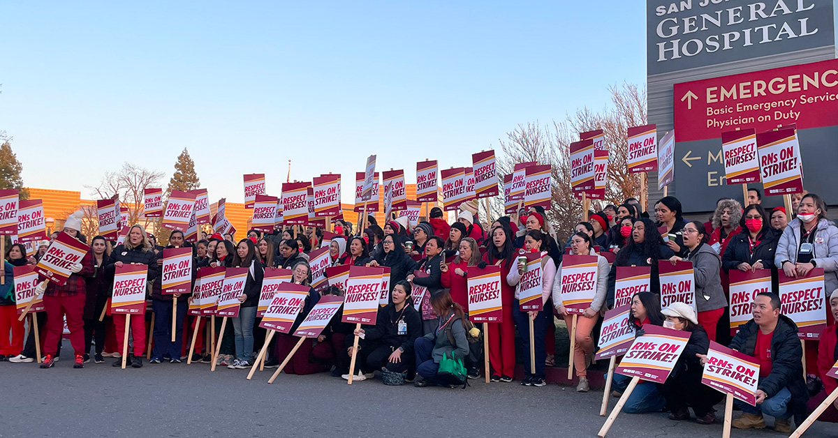 Large group of nurses outside San Joquin General Hospital, smiling and holding signs "RNs On Strike" and "Respect Nurses"