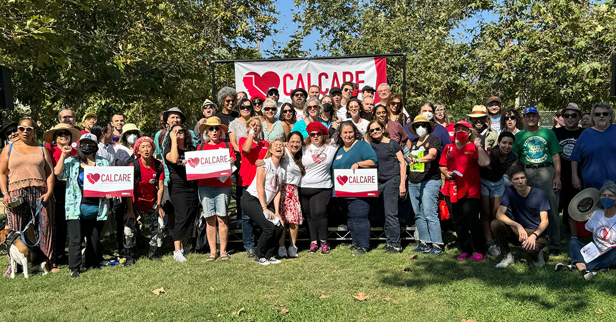 Large group of people in park standing and sitting in front of CalCare banner, some holding CalCare signs