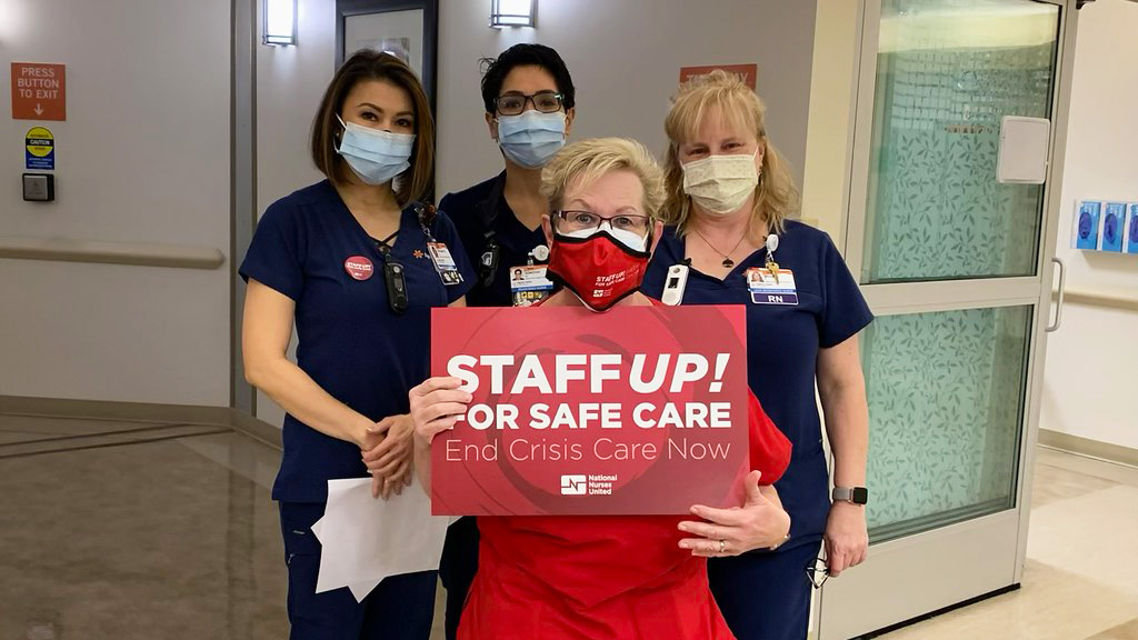 Four nurses inside hospital, one holds sign "Staff Up for Safe Patient Care"