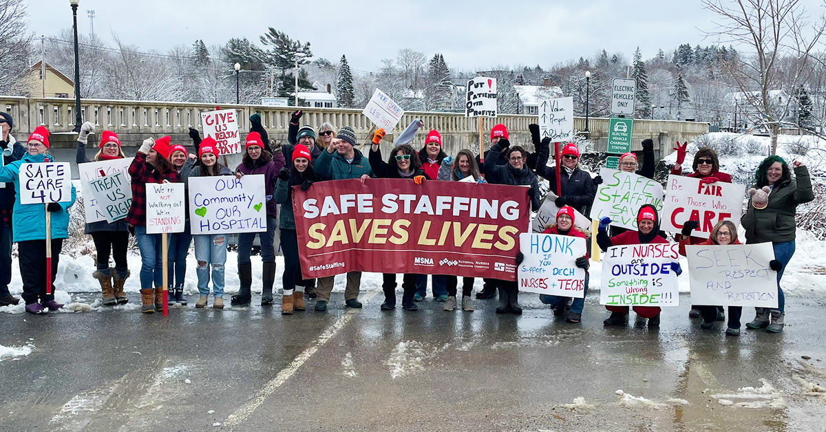 Large group of nurses outside in snow, holding banner "Safe Staffing Saves Lives" and other signs calling for improvements in the workplace.