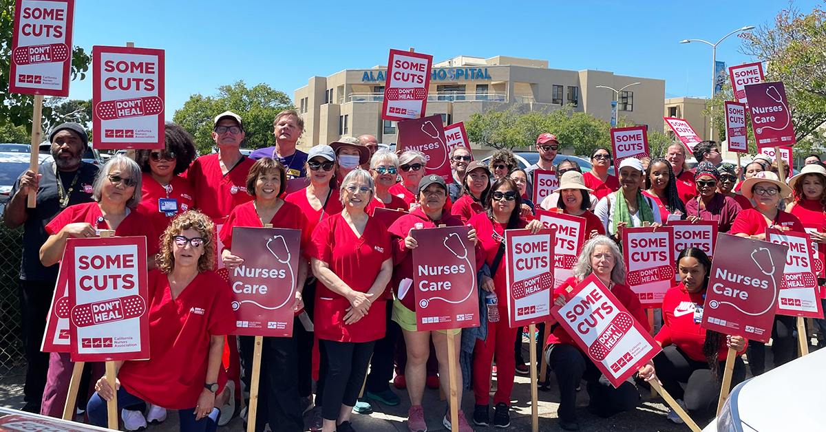 Alameda RNs in front of hospital holding signs "Some cuts don't heal" and "Nurses care"