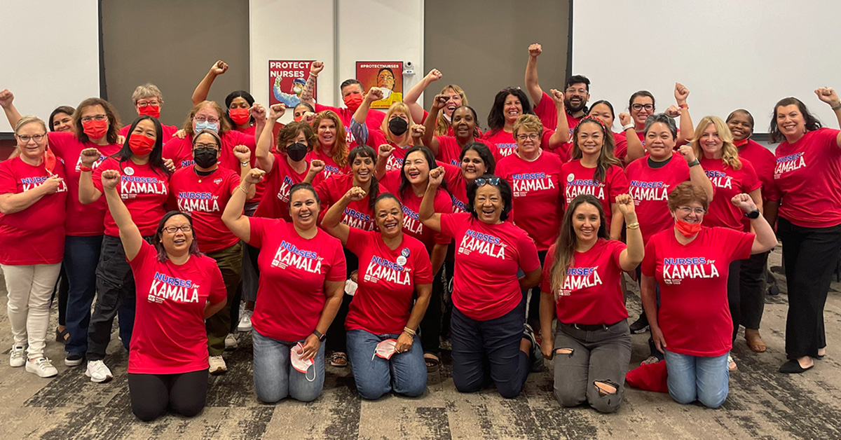 Large group of nurses indoors wearing red "Nurses for Kamala" signs with raised fists