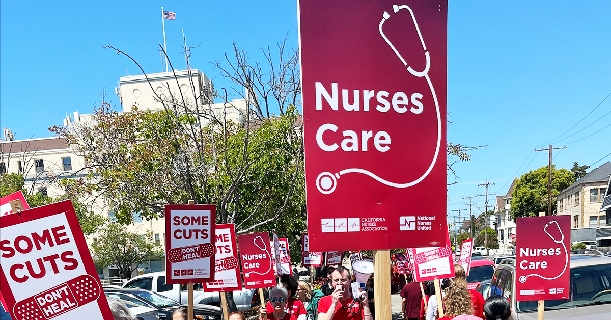 Marching on sidewalk with signs "Nurses care" (stethoscope image) and "Some cuts don't heal" (bandaid image)