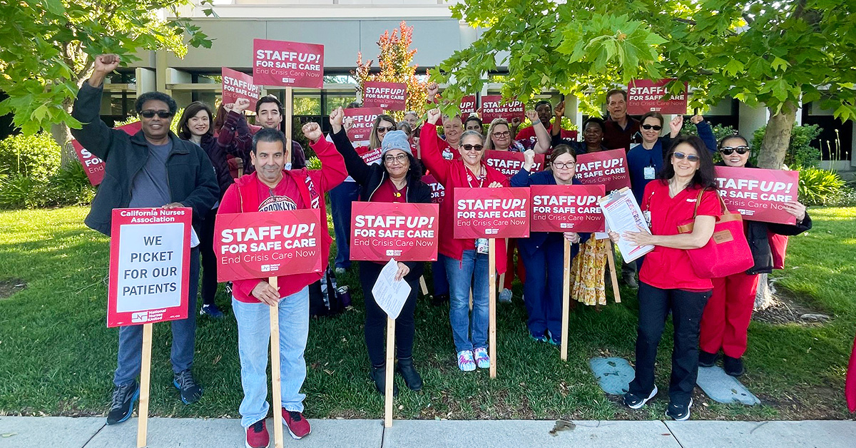 Large group of nurses standing outside hospital with raised fists, holding signs "Staff Up for Safe Patient Care"