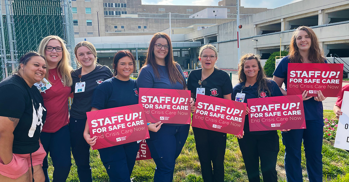 Group of nurses outside hospital smiling, holding signs "Staff Up for Safe Patient Care"