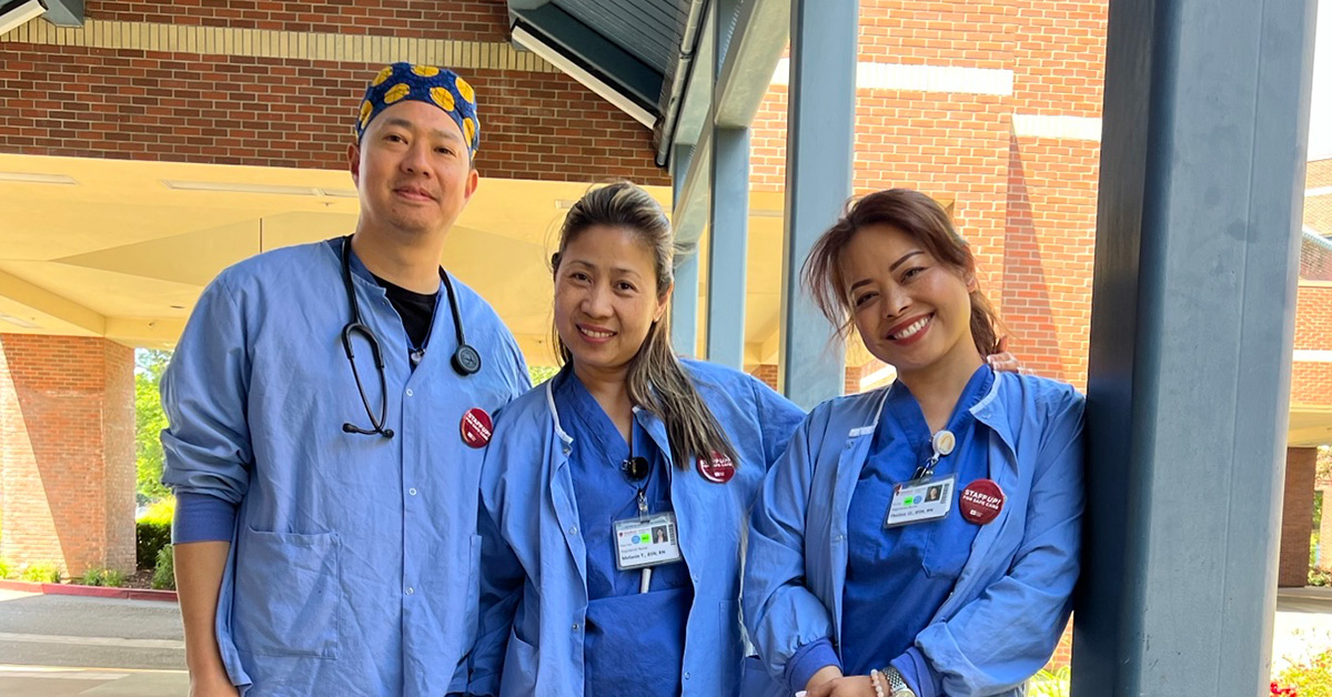 3 nurses in blue scrubs outside standing side-by-side, smiling