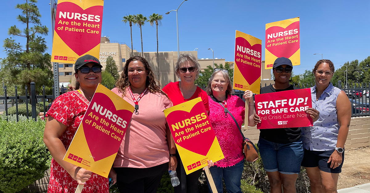 Group of six nurses outside hospital, smiling, holding signs "Nurses are the hear of patient care"