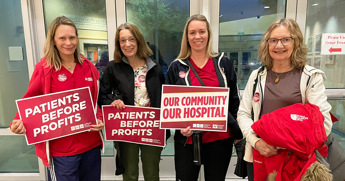 Four nurses inside hospital, smiling, holding signs "Patients Before Profits" and "Our Community, Our Hospital"