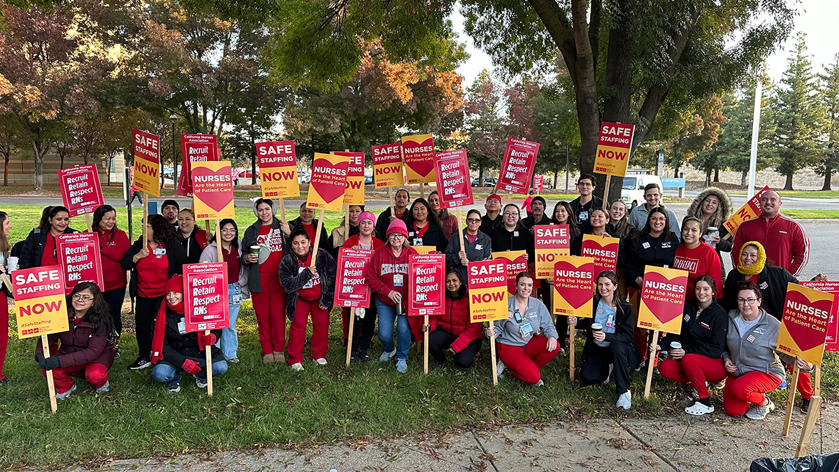 Large group of nurses outside holding signs "Safe Staffing NOW", "Recruit, Retain, Respect RNs", and "Nurses are the heart of patient care"