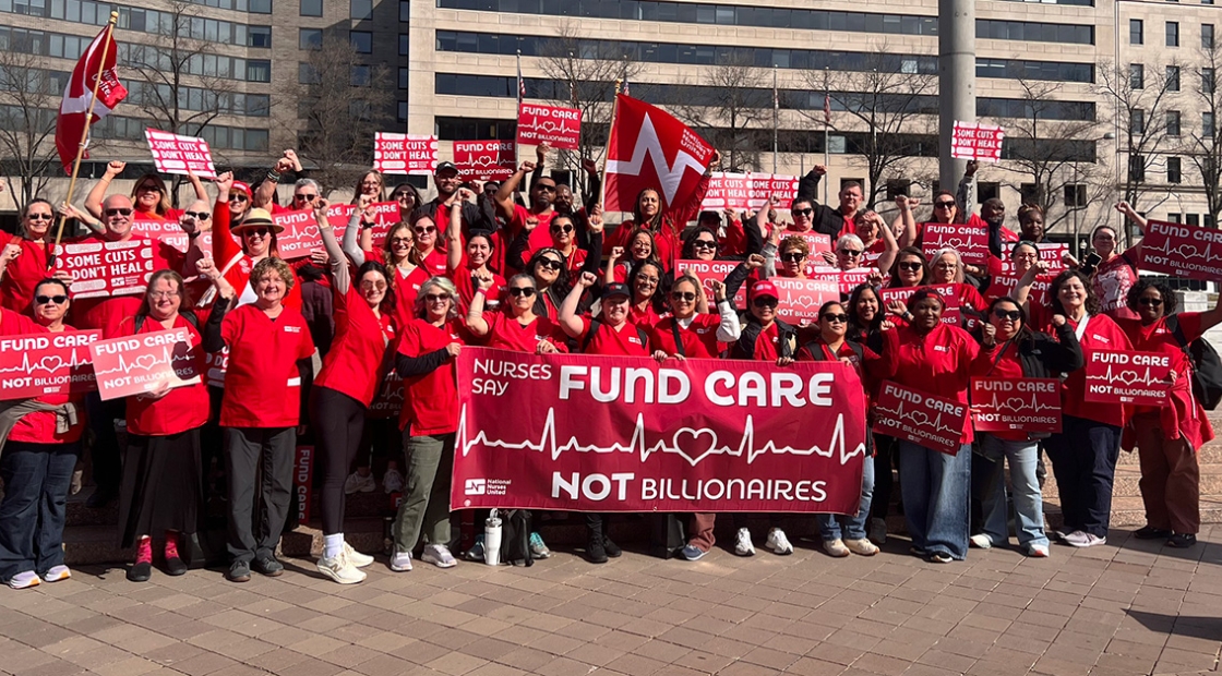 Large group of nurses outside holding banner and signs "Nurses Say: Fund Care Not Billionaires"