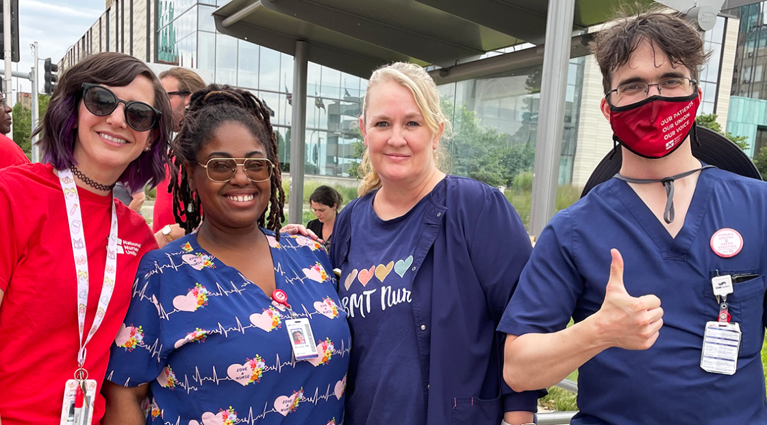 Four SLU Hospital nurses smiling in front of hospital, one giving a thumbs up.