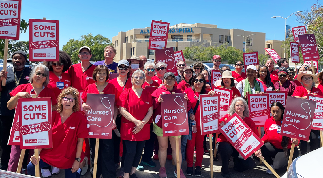 Alameda RNs in front of hospital holding signs "Some cuts don't heal" and "Nurses care"