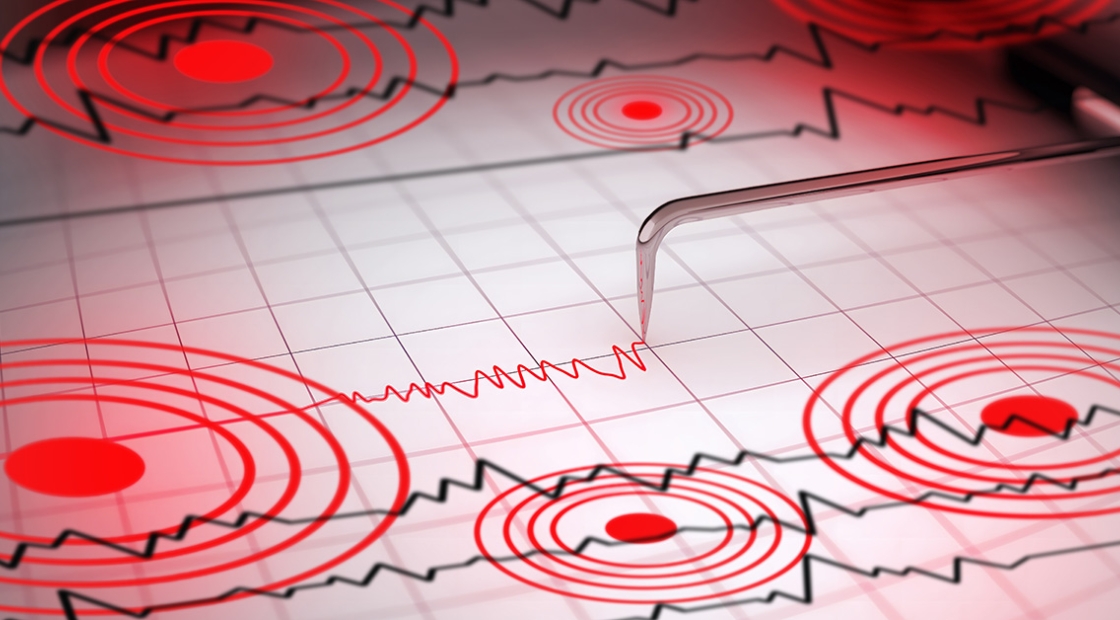 Close-up of a seismograph machine needle tracing seismic waves, indicative of earthquake activity, on graph paper with epicenter rings.