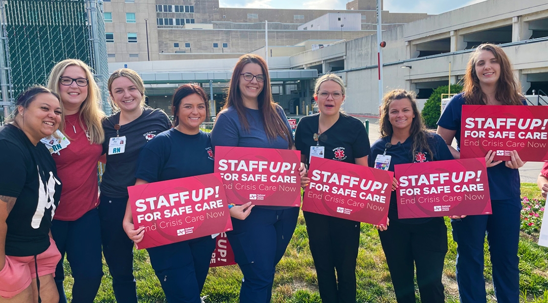 Group of nurses outside hospital smiling, holding signs "Staff Up for Safe Patient Care"