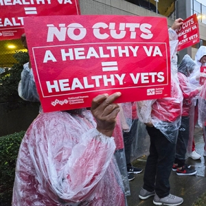 Nurses on picket line outside hospital in the rain. One holds sign "No Cuts: A Healthy VA = Healthy Vets