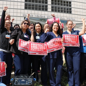 Group of nurses with raised fists, holding signs "Protect Nurses, Patients, Public Health"