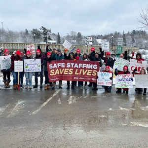 Large group of nurses outside in snow, holding banner "Safe Staffing Saves Lives" and other signs calling for improvements in the workplace.