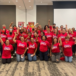 Large group of nurses indoors wearing red "Nurses for Kamala" signs with raised fists