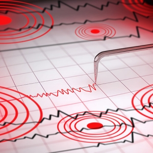 Close-up of a seismograph machine needle tracing seismic waves, indicative of earthquake activity, on graph paper with epicenter rings.