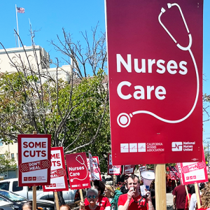 Marching on sidewalk with signs "Nurses care" (stethoscope image) and "Some cuts don't heal" (bandaid image)