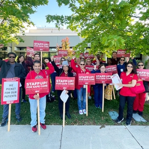 Large group of nurses standing outside hospital with raised fists, holding signs "Staff Up for Safe Patient Care"