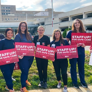 Group of nurses outside hospital smiling, holding signs "Staff Up for Safe Patient Care"