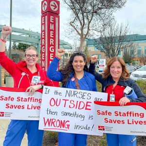 Three nureses outside of hospital, smiling, fists raised, and holding signs "Safe Staffing Saves Lives"