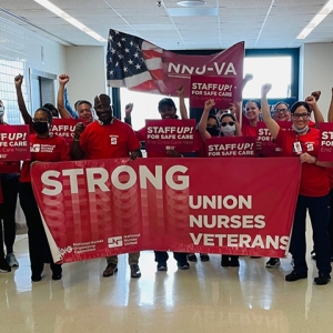 Larg group of nurses inside hospital wearing red, fists raised, holding banner "Strong Union, Strong Nurses, Strong Veterans"