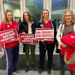 Four nurses inside hospital, smiling, holding signs "Patients Before Profits" and "Our Community, Our Hospital"