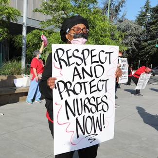 Nurse outside facility with sign "Respect and protect nurses now!"